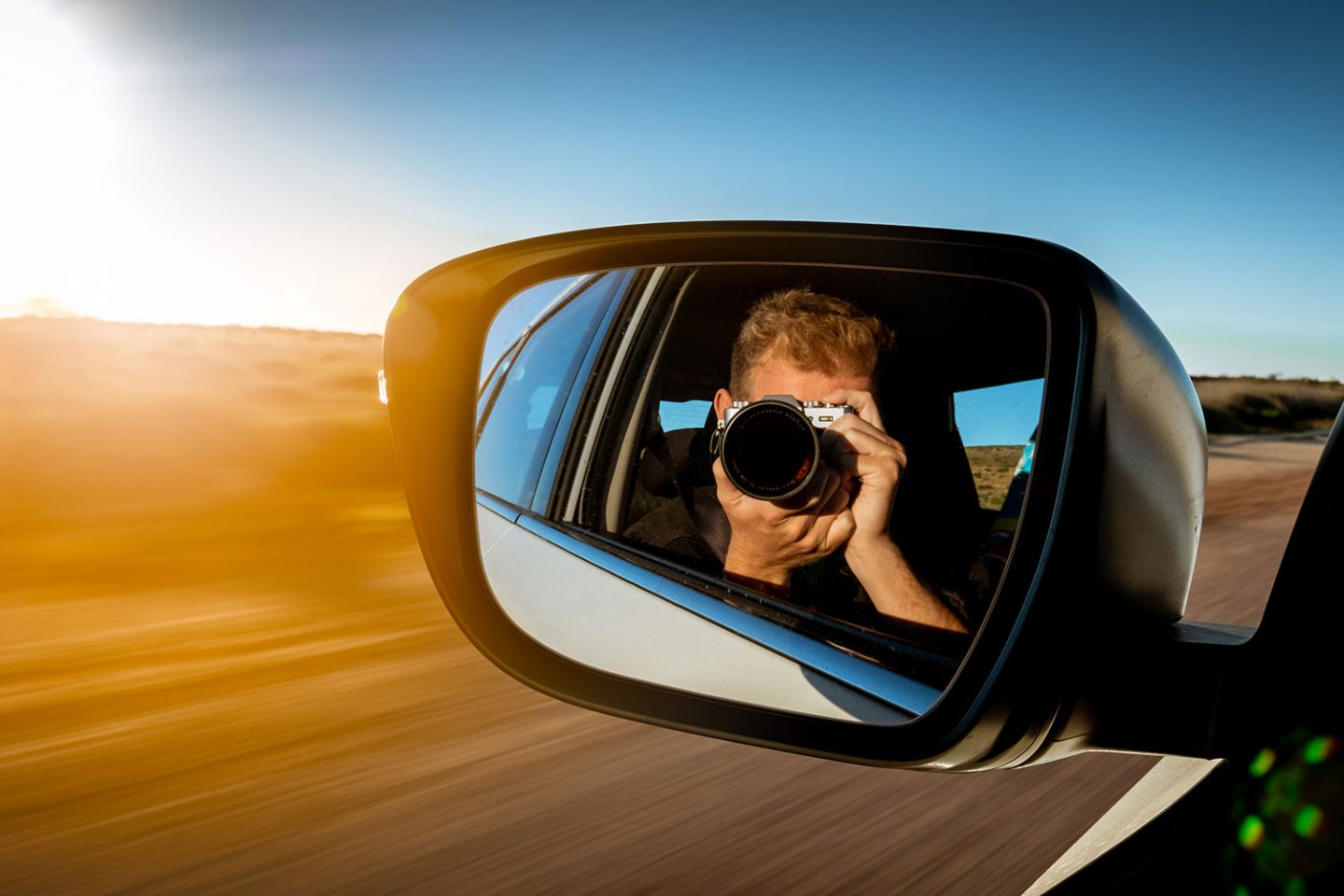 Man taking a photo of himself in a car mirror as car is driving with the warm bright light of the sunset hits the car mirror