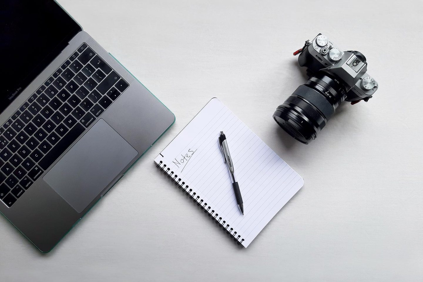 A camera, notepad and laptop arranged on top of a white desk