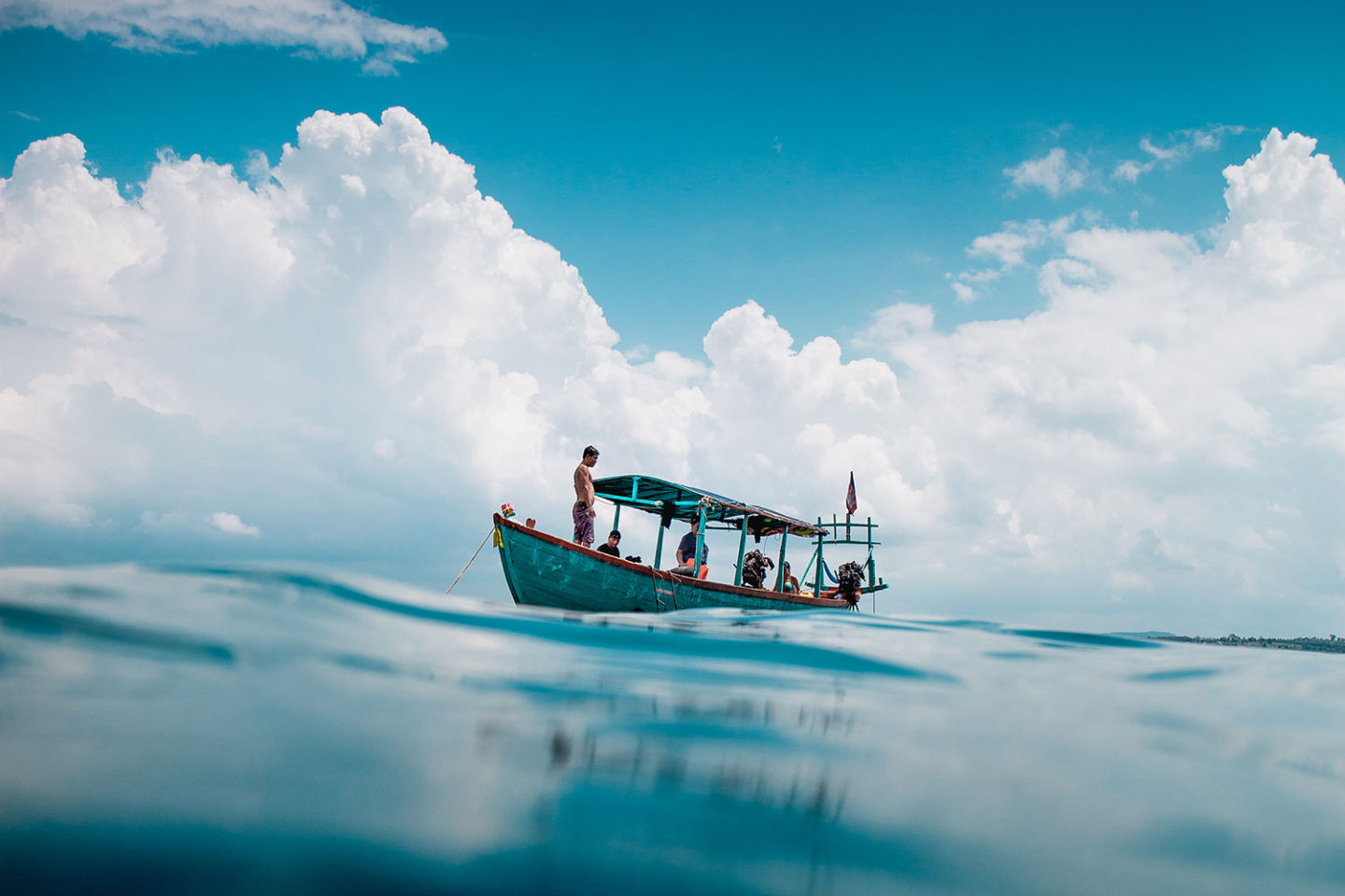 A man stands at the front of a boat with beautiful crystal clear water in the foreground and a dramatic looking sky behind.