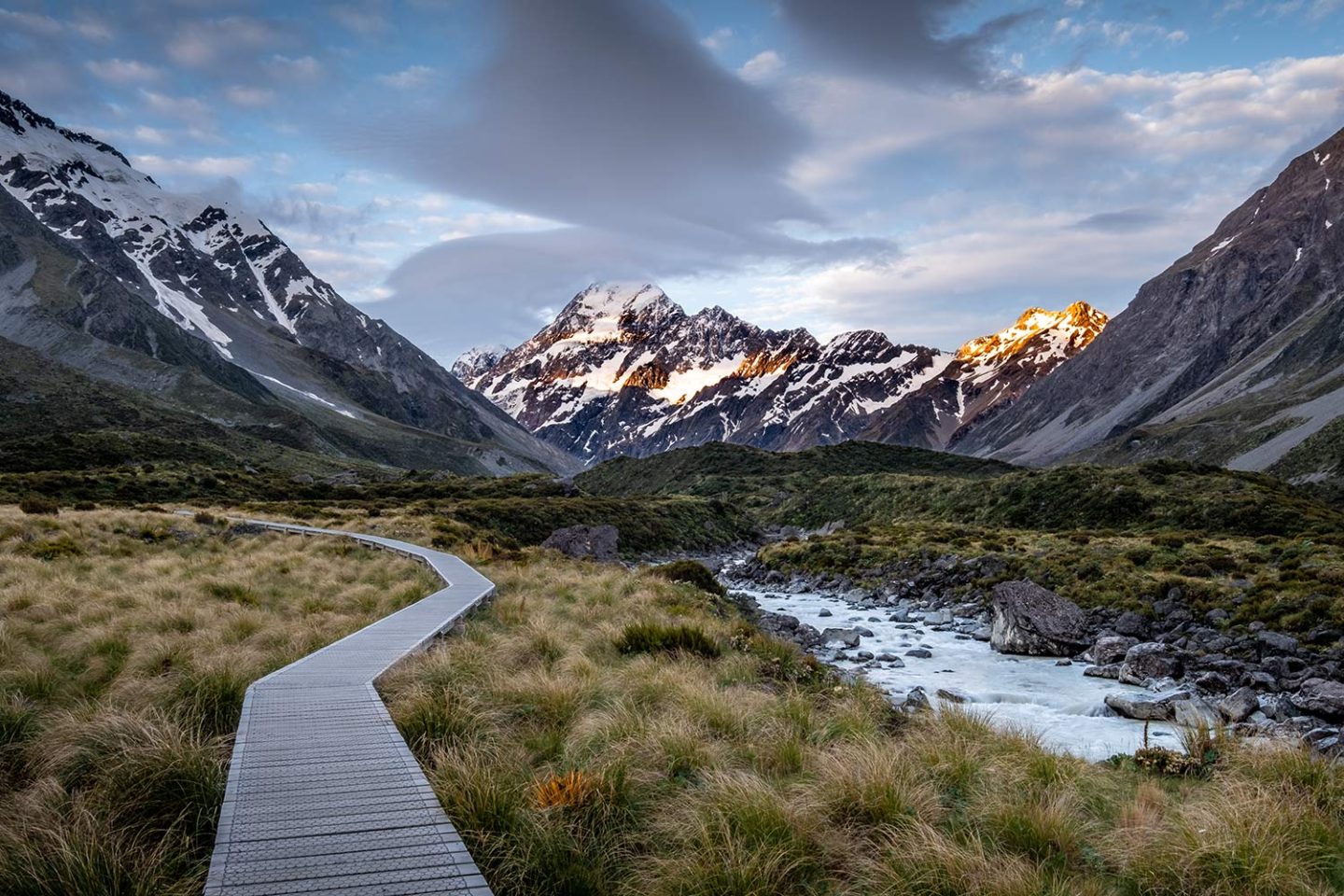 A wooden path leads towards mountains through long grass.