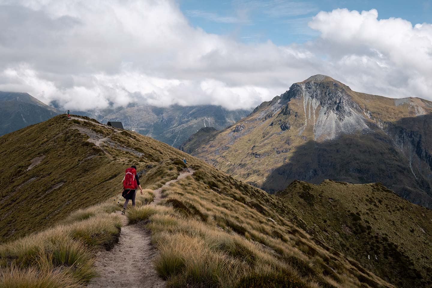A woman with a large red backpack walks along a ridge line on the Kepler Track, in front of her are large mountains.
