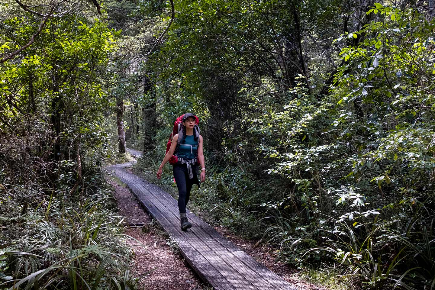 A women walking along a wooden path with a red hiking pack on her back. She is surrounded by lots of lush green forest.