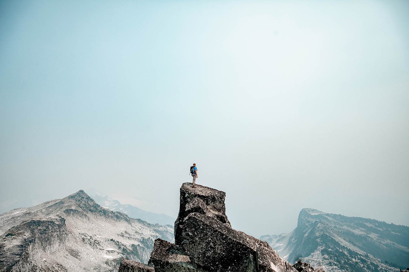 A man stands on the top of a large rock and looks at the snow tipped mountain range in front of him.